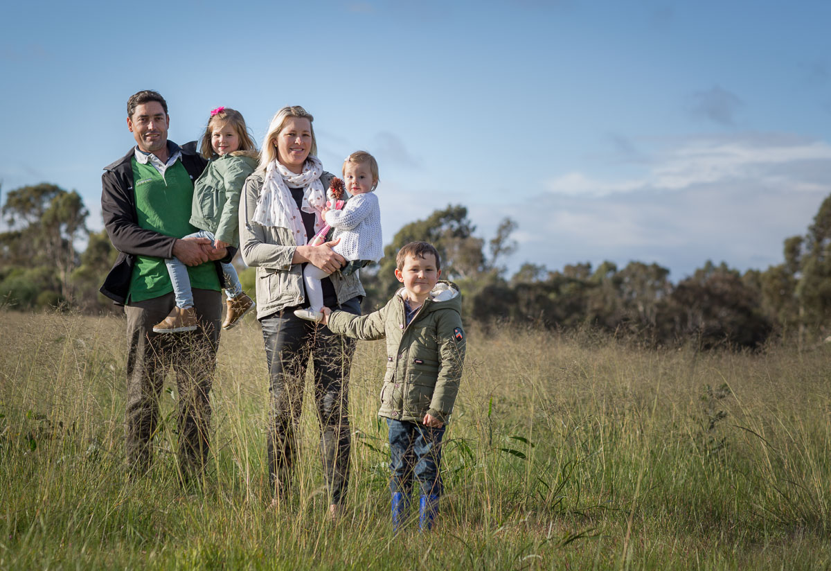 Photo off attractive young couple with their three children standing in a grassy field. Photo by Caro Telfer, photographer.
