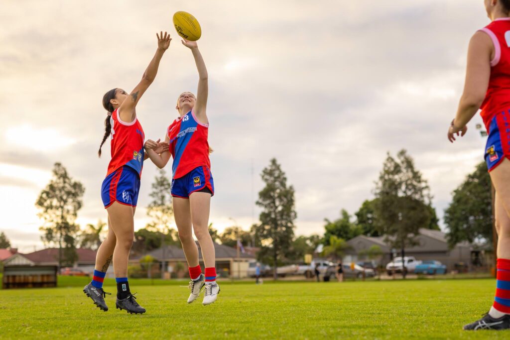 Two young women playing Aussie Rules football jump to tap the ball.