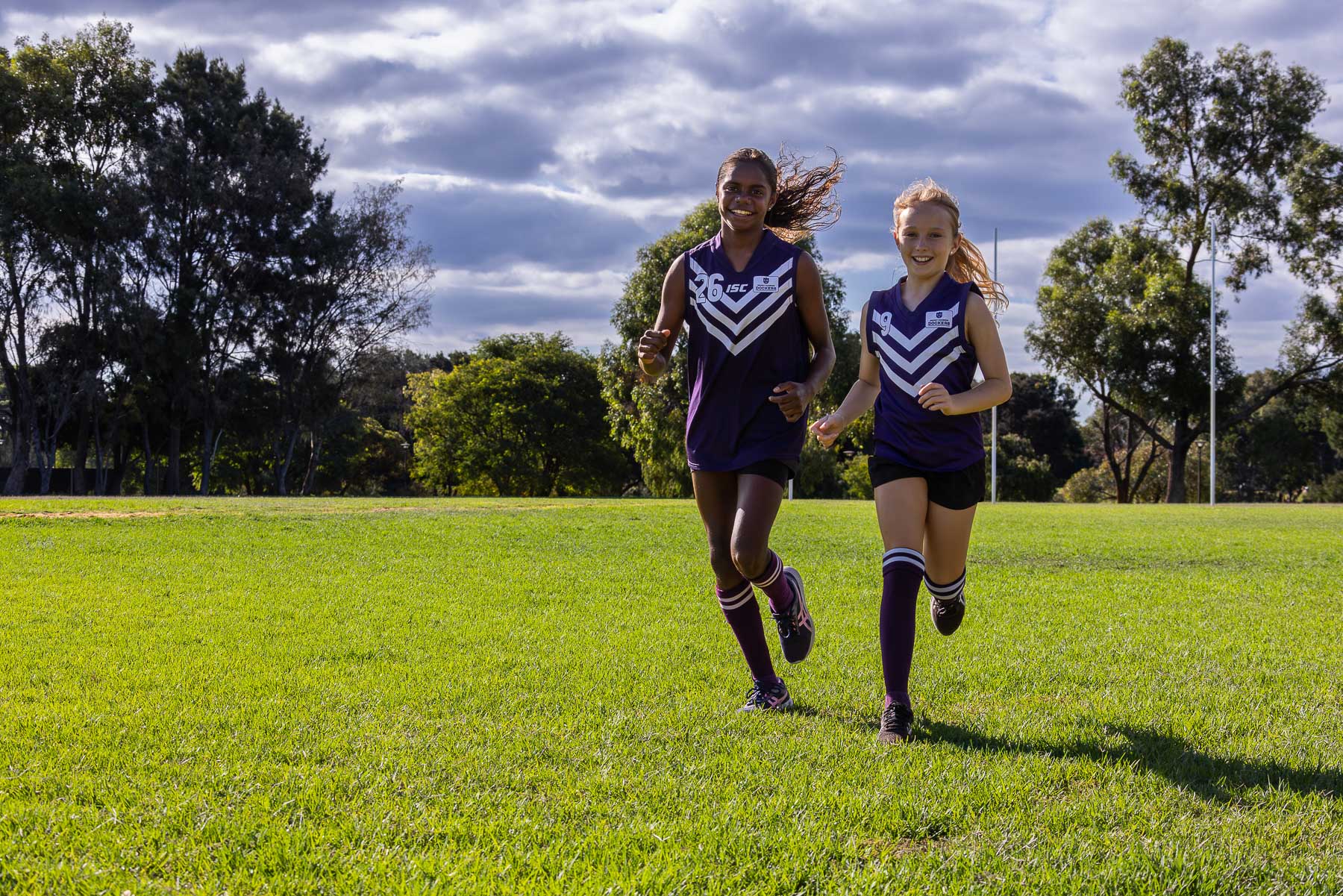two girls in footy uniforms jogging on green grass playing surface