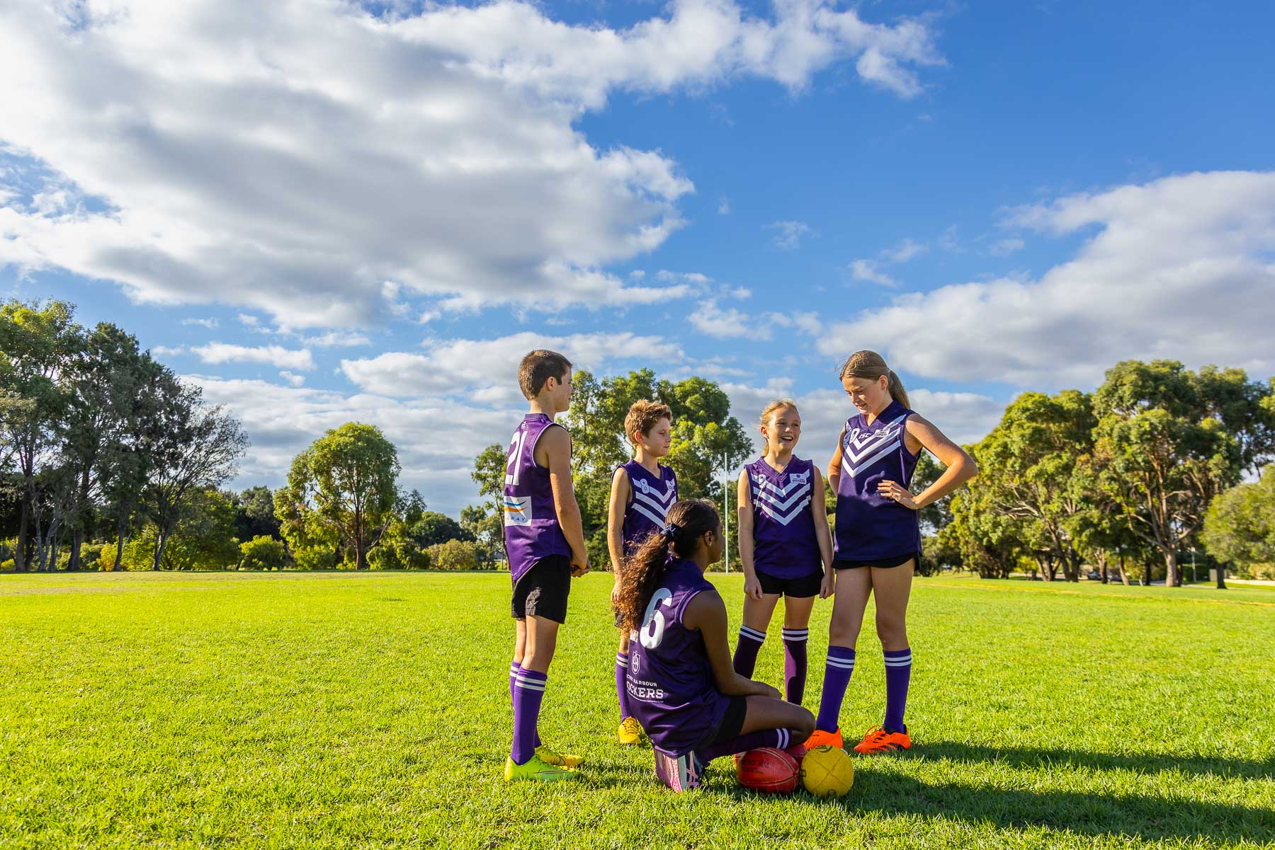 group of five schoolchildren in football uniforms standing and sitting around on football oval.