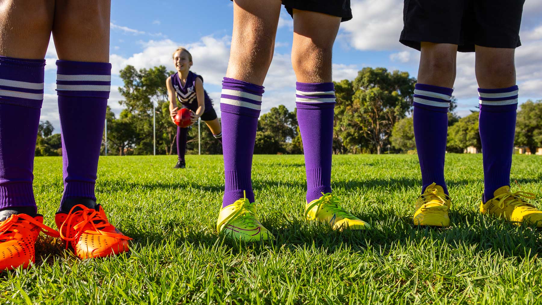 image with three pairs of legs wearing footy boots and long purple sock with one child behind handling AFL football