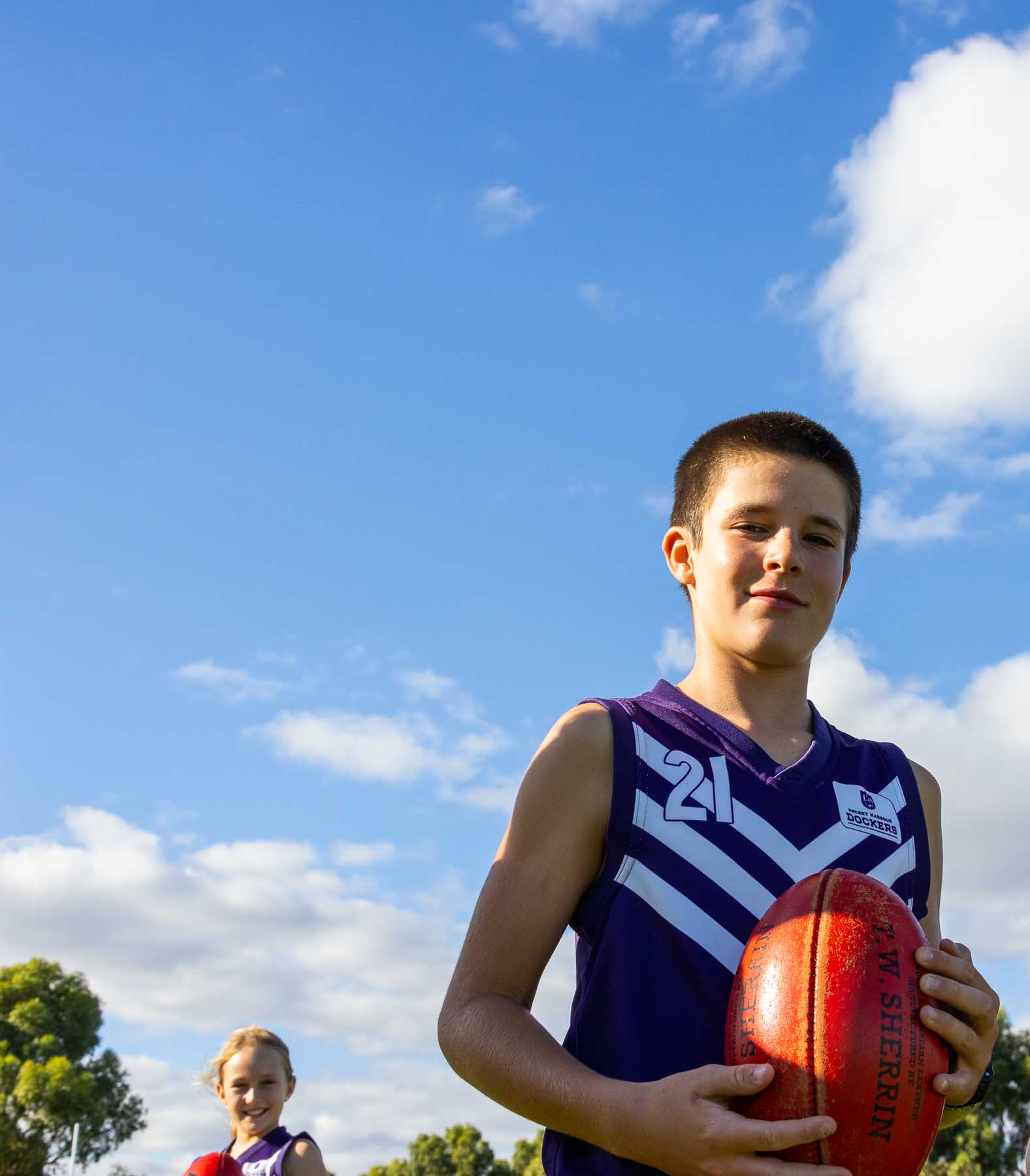 one boy holding an AFL football with teammate small in background