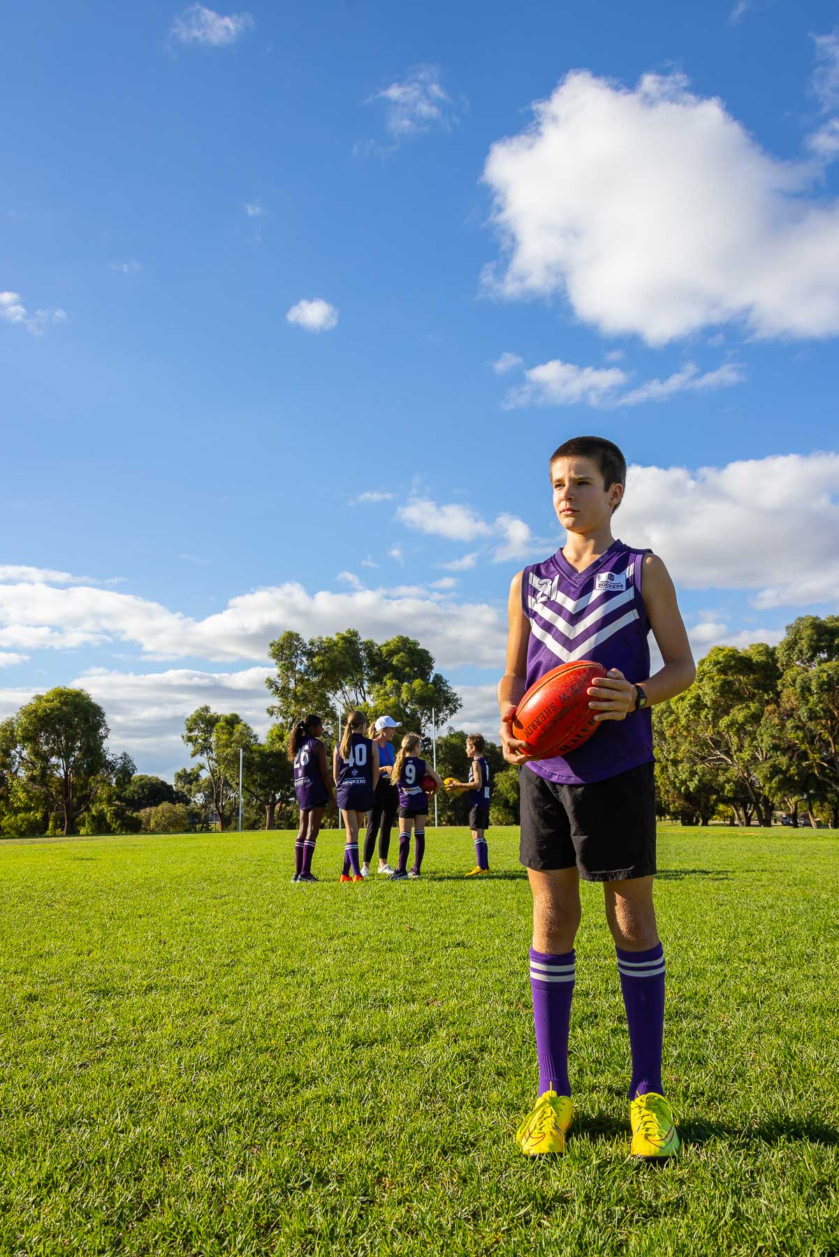 vertical image of one boy holding a red AFL football with teammates small in background