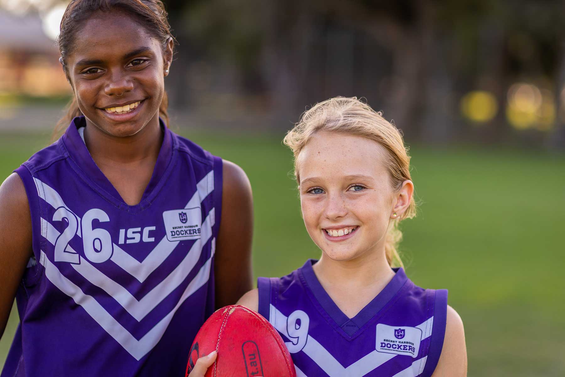 head and shoulders of two smiling schoolgirls looking at the camera holding an AFL football.