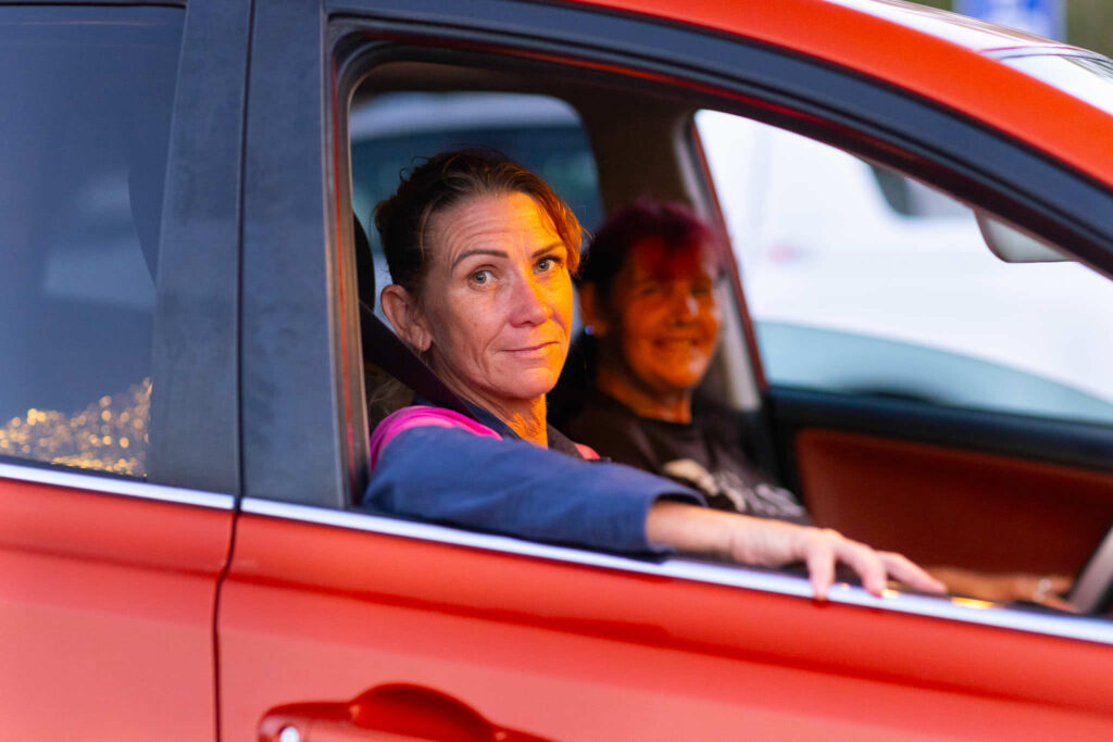 Photo of two women sitting in a red car and the driver is looking out the open window with her arm resting on the opening.