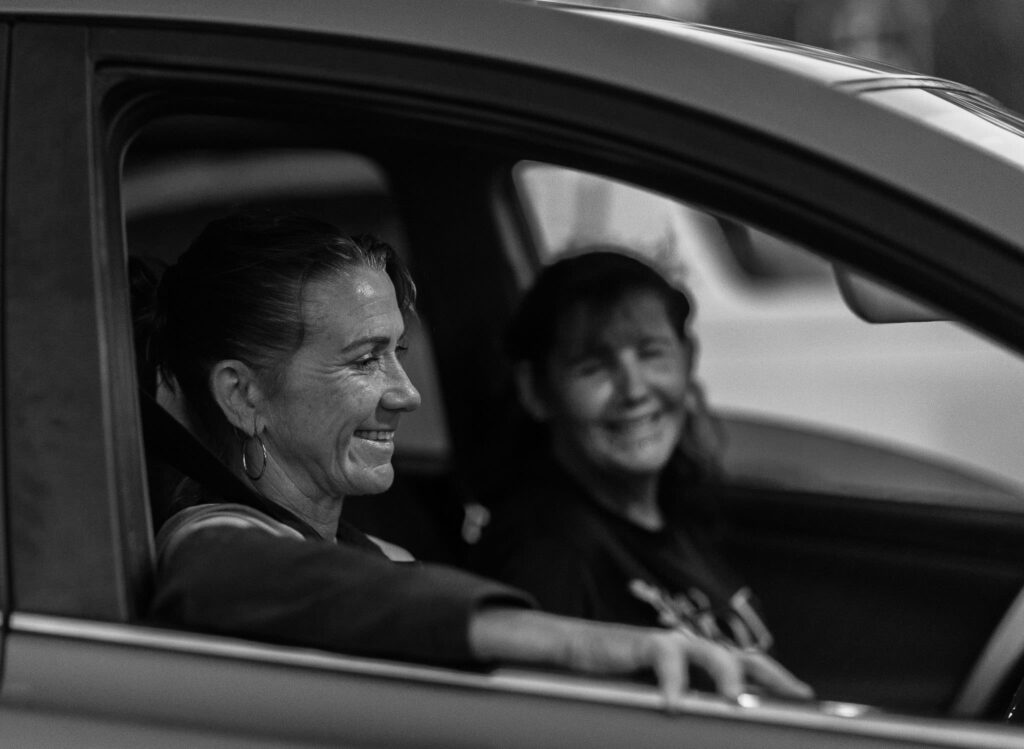 A dark black and white image of two women sitting in a car with the driver's arm resting on the window ledge.