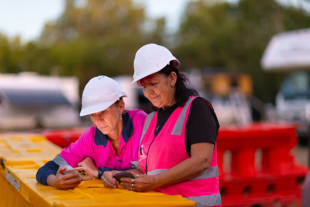 photo of two women wearing pink hi-vis and white hard-hats looking at their phones and leaning on a yellow barrier.