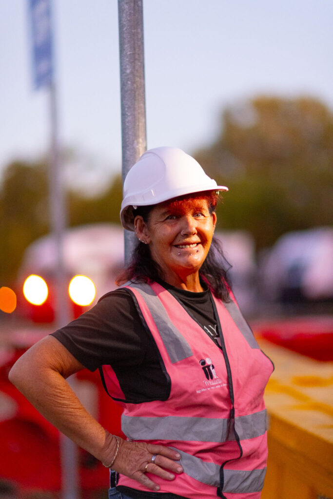 Photo of a lady in her 50s wearing a hard-had and pink hi-vis vest. She has one hand on her hip and is looking at the camera, showing facial piercings.