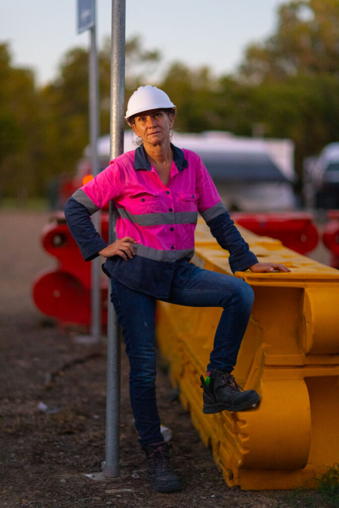 One female worker wearing hi-vis and denim with a white hard-had. She is standing with one foot up on a yellow safety barrier.